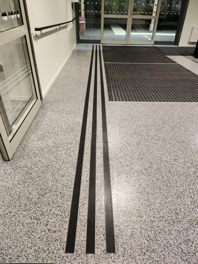 Svart aluminiumlist med halksäkert ilägg på ljust stengolv – leder in och ut från entrén.